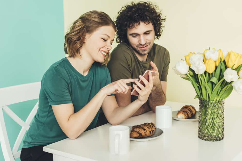 A couple eats croissants and coffee while sitting on a table. The woman holds a phone while he looks at what she's pointing at.