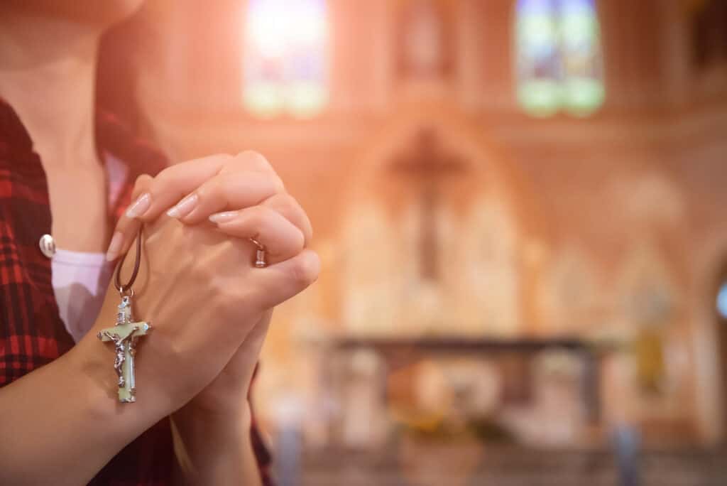 Close up of a woman holding a rosary against cross and praying at a church.