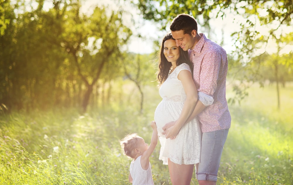 A man's arms are around his wife's pregnant belly with another child standing in front of them.