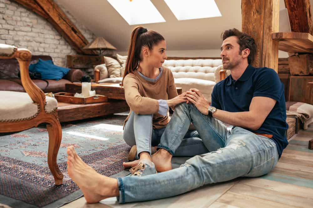 A man and woman sit on the floor of a living room while having a serious talk.