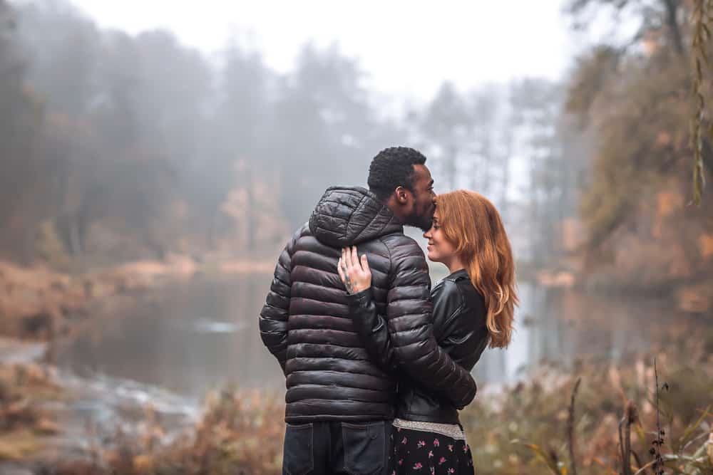A man kisses a woman's forehead in a foggy lake landscape.