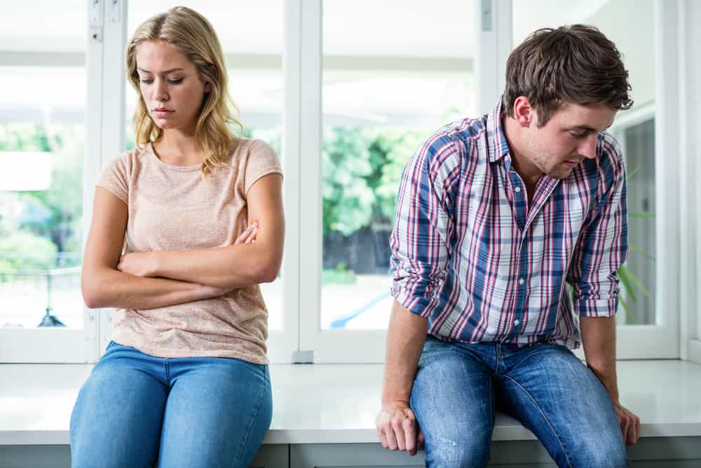 A couple sits angrily next to one another by a window.