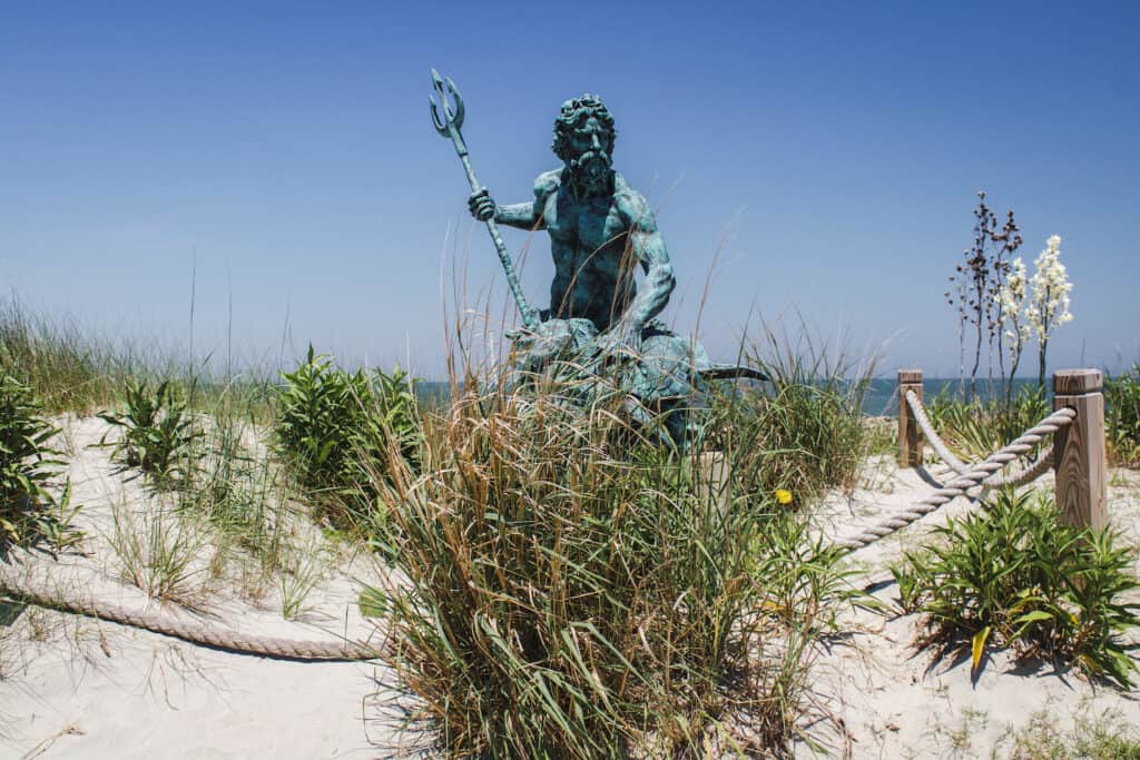 A statue sits on the beach on a sunny day surrounded by grass with the ocean behind.