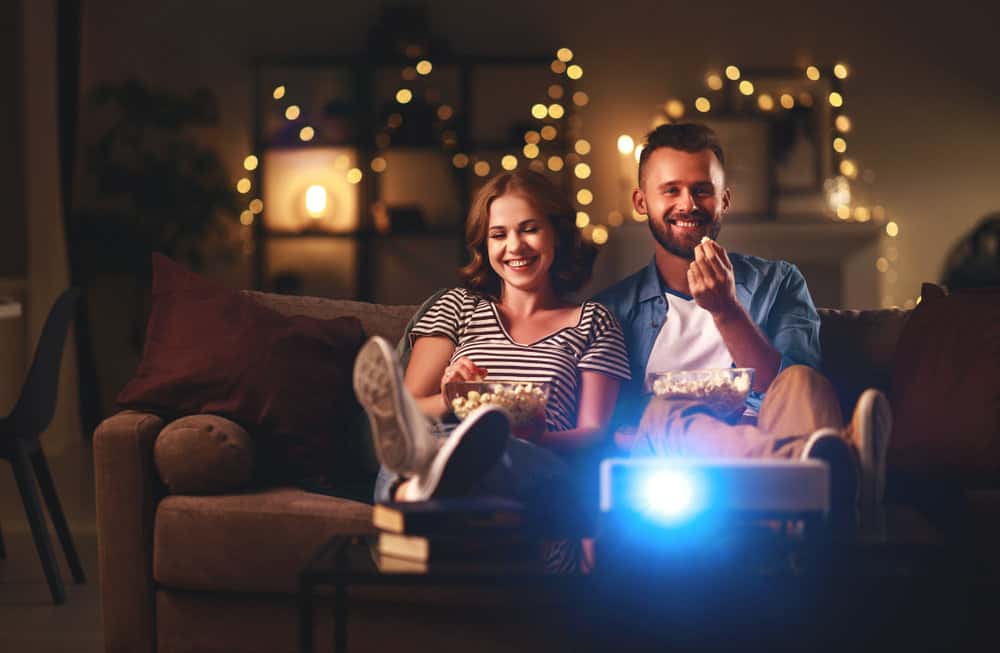 A couple sitting together eating from small bowls of popcorn with a projector placed on the table in front of them. 