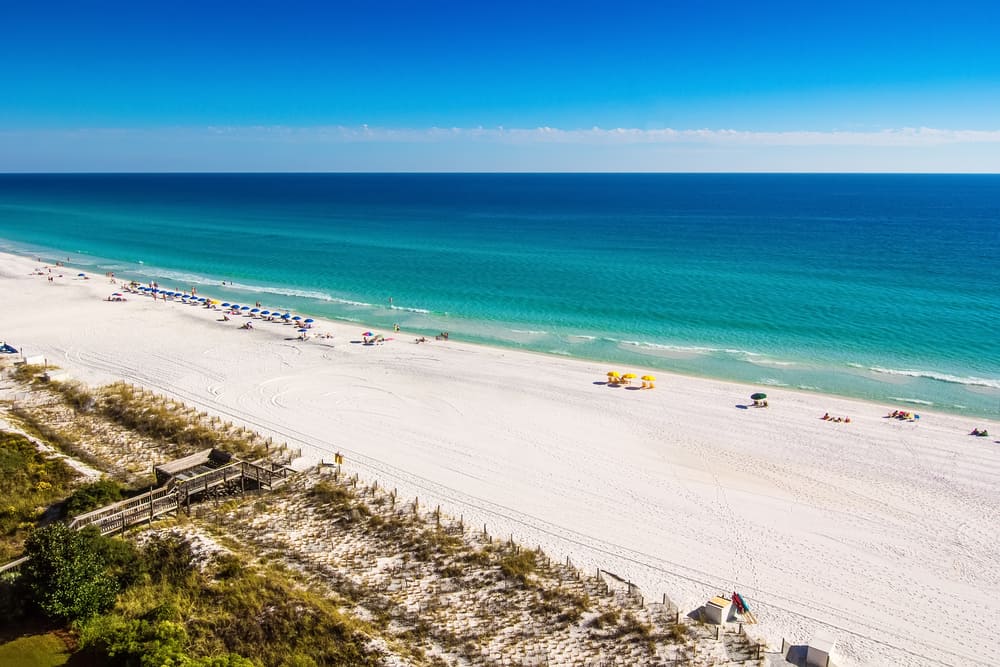 A white sand beach with people laying out in front of bright blue water.