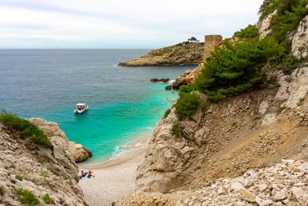 A boat floating on the water near the shoreline. People are sitting on the sand of the beach.