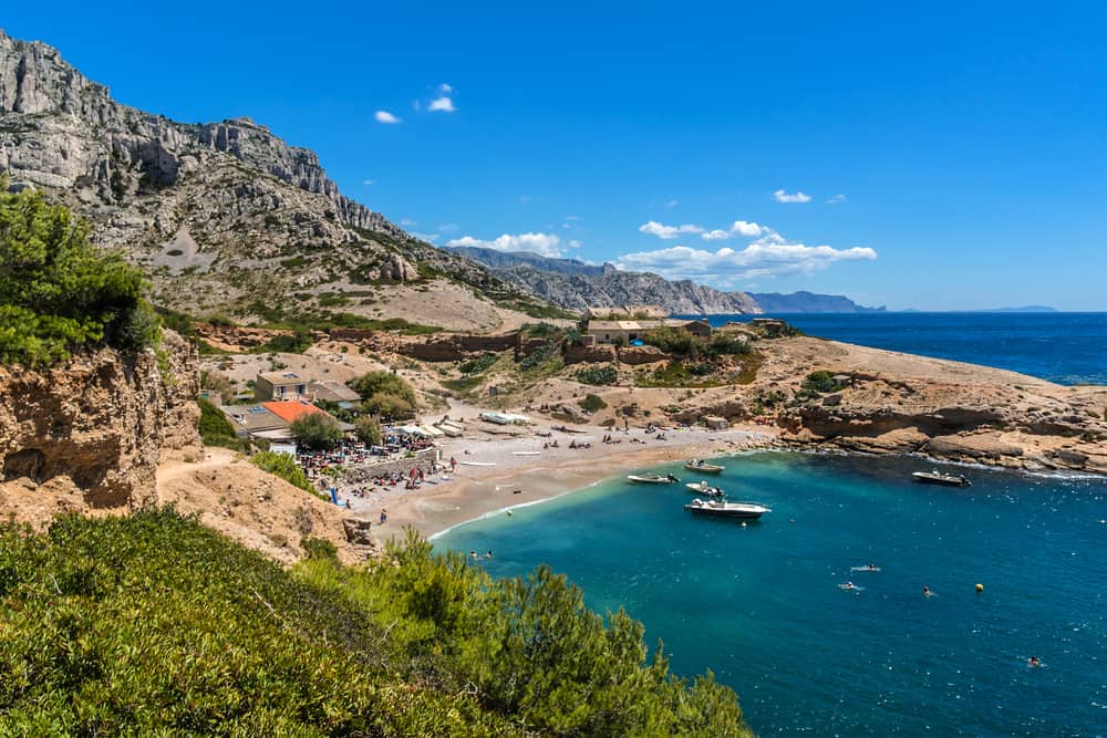 Boats floating on the blue ocean by an island with a tall mountain under a blue sky. People are mingling on the beach.